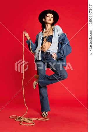 Rodeo. Portrait of beautiful young woman in jeans clothes and black hat posing with string like cowboy against red studio background. Rodeo. Portrait of beautiful young woman in jeans clothes and black hat posing with string like cowboy against red studio background. 100547547
