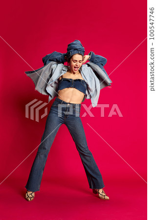 Positivity. Music. Full-length portrait of young beautiful woman in stylish jeans clothes posing against red studio background. Concept of emotions and fashion Positivity. Music. Full-length portrait of young beautiful woman in stylish jeans clothes posing against red studio background. Concept of emotions and fashion 100547746