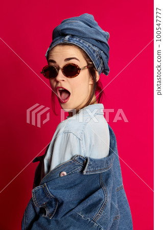 Shocked and excited face. Young beautiful woman posing in trendy stylish jeans clothes withsunglasses against red studio background. Concept of emotions and fashion 100547777