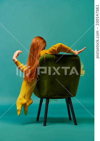 Creative portrait of unknown redhead girl with long straight silky hair posing on armchair, sitting in strange poses over cyan color background. Impersonal emotions, body language 100547861