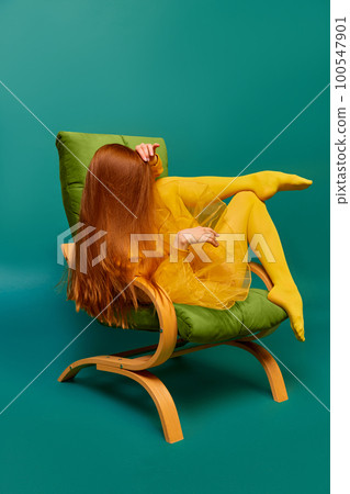 Creative portrait of unknown redhead girl with long straight silky hair posing on armchair, sitting in strange poses over cyan color background. Impersonal emotions, body language 100547901