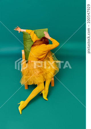Creative portrait of unknown redhead girl with long straight silky hair posing on armchair, sitting in strange poses over cyan color background. Impersonal emotions, body language 100547929