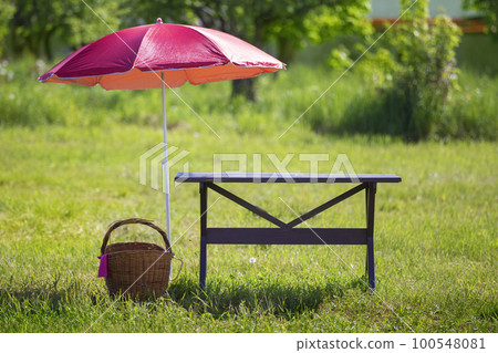 For a picnic. On a green meadow there is a wooden table, a wicker basket and zones from the sun. 100548081