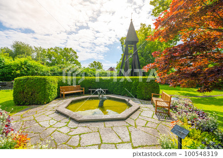 A small stone fountain with a statue and a wooden bell tower on Galarvarvskyrkogarden Cemetery in Stockholm, Sweden, on a sunny day 100548319