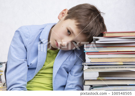 A tired school-age boy sits between stacks of books. Sad child with books. 100548505