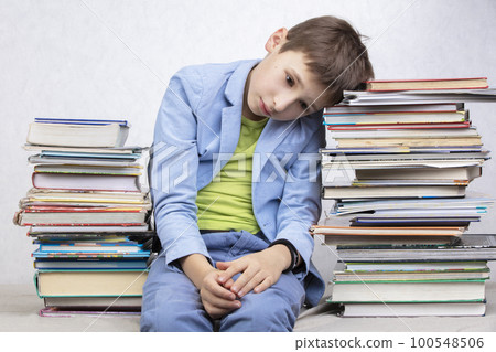 A tired school-age boy sits between stacks of books. Sad child with books. 100548506