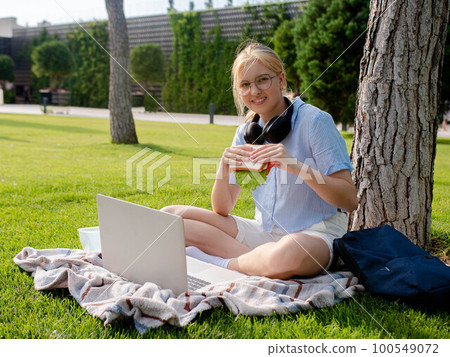 A young student girl in glasses sits in a park on the grass, uses a laptop, eats a sandwich. A young student girl in glasses sits in a park on the grass, uses a laptop, eats a sandwich. 100549072