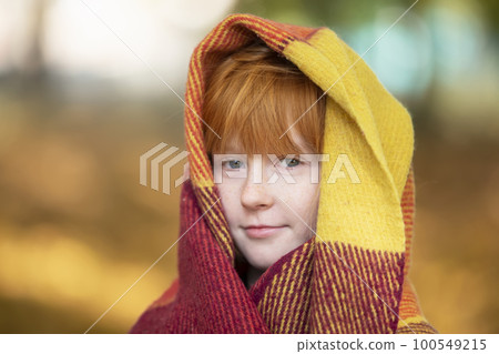 A red-haired girl wrapped herself in a woolen blanket in an autumn park. Child in autumn. 100549215