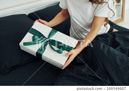 Gift box in hands of young woman. Interior and design of beautiful modern bedroom at daytime Gift box in hands of young woman. Interior and design of beautiful modern bedroom at daytime 100549887
