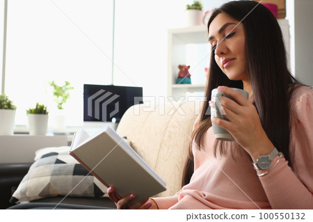 Young woman is reading book and holding cup in living room on sofa 100550132