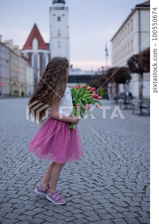 Adorable 9 years old girl with long curly hair walking through central square of the town Olesnica, Poland. Beautiful girl walking with bouquet of tulip flowers through spring European city. 100550674