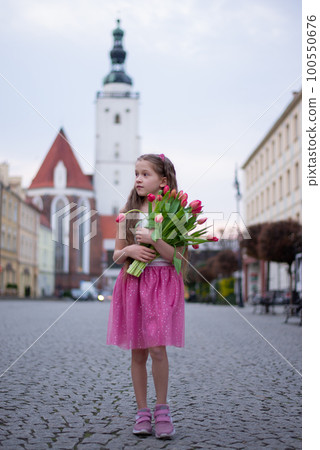 Adorable 9 years old girl with long curly hair walking through central square of the town Olesnica, Poland. Beautiful girl walking with bouquet of tulip flowers through spring European city. Adorable 9 years old girl with long curly hair walking through central square of the town Olesnica, Poland. Beautiful girl walking with bouquet of tulip flowers through spring European city. 100550676