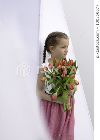 Adorable 9 years old girl with long curly hair walking through central square of the town Olesnica, Poland. Beautiful girl walking with bouquet of tulip flowers through spring European city. 100550677