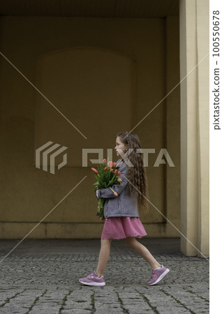 Adorable 9 years old girl with long curly hair walking through central square of the town Olesnica, Poland. Beautiful girl walking with bouquet of tulip flowers through spring European city. Adorable 9 years old girl with long curly hair walking through central square of the town Olesnica, Poland. Beautiful girl walking with bouquet of tulip flowers through spring European city. 100550678
