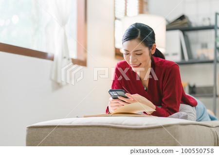 In a cozy living room at home, a beautiful young woman laying on the sofa, engrossed in messaging on her smartphone with deft fingers. 100550758