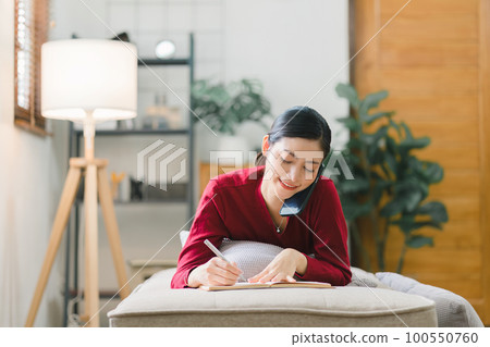A beautiful businesswoman is speaking on her smartphone while working and writing notes while lying on the couch in the living room. 100550760