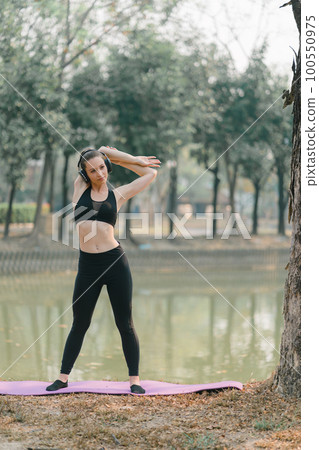 In an Autumn city park background, a stunning sportswoman in sportswear is smiling while stretching her body and performing flexibility exercises as a warm-up before a running workout. 100550975