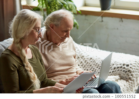 Serious concentrated senior people, retired couple, man and woman sitting on couch at home and checking documents, looking on laptop 100551552