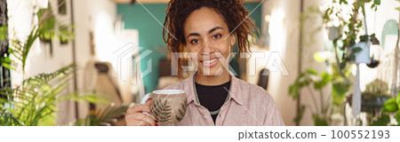 Smiling woman floristic studio owner standing with cup of tea and laptop during break Smiling woman floristic studio owner standing with cup of tea and laptop during break 100552193