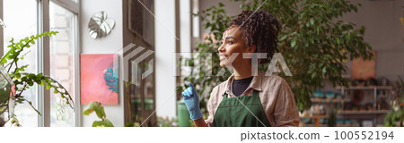 Attractive smiling florist stands near windows on background of her own floral decor studio Attractive smiling florist stands near windows on background of her own floral decor studio 100552194