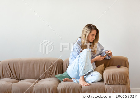 Digital addiction - woman sitting at home on a couch, lost in the endless scroll of her smartphone screen. 100553027
