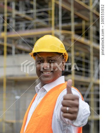 Smiling Indian construction worker wearing safety helmet and vest posing on construction site showing thumbs up gesture 100555852