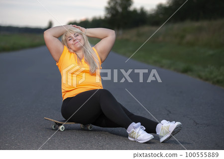 Chubby middle aged European woman, overweight, in a tracksuit with a skateboard, tired after playing sports, resting. 100555889