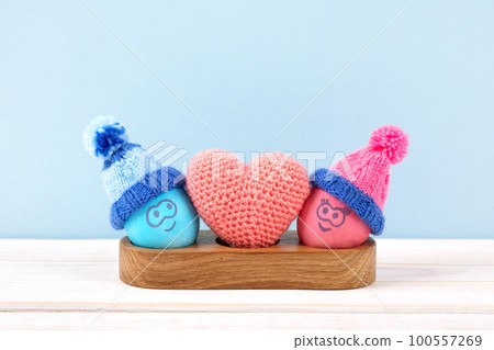 Two Easter eggs with funny faces and crocheted hats in wooden egg stand and a crocheted heart on a wooden table. Happy Easter concept. Greeting card. 100557269
