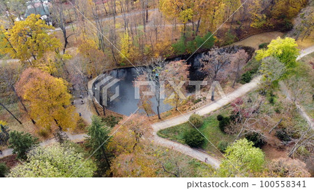 Flying over the autumn park. Many trees with yellow green and fallen leaves, lakes, people walking along dirt paths in park on autumn day. Top view. Aerial drone view. Beautiful natural background Flying over the autumn park. Many trees with yellow green and fallen leaves, lakes, people walking along dirt paths in park on autumn day. Top view. Aerial drone view. Beautiful natural background 100558341