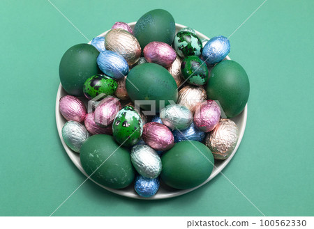 Delicious Easter food, treat. Colored chicken and quail eggs, chocolate eggs, sweets wrapped in bright foil on plate, flatly top view. Close up, horizontal. Christian holiday tradition. Confectionary Delicious Easter food, treat. Colored chicken and quail eggs, chocolate eggs, sweets wrapped in bright foil on plate, flatly top view. Close up, horizontal. Christian holiday tradition. Confectionary 100562330