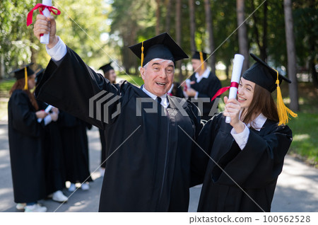 A group of graduates in robes outdoors. An elderly man and a young woman congratulate each other on their graduation. 100562528