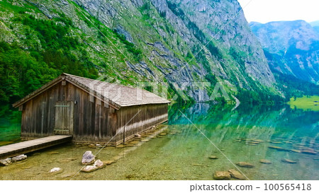 Konigssee, Germany - June 16, 2018: side view of lakeside cabin or traditional wooden boat house in Obersee on the left with a peaceful and clear lake on a sunny summer day 100565418