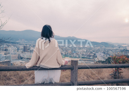 照片中一位女士拿著咖啡杯站在早晨的陽光下，背景是城市 100567596