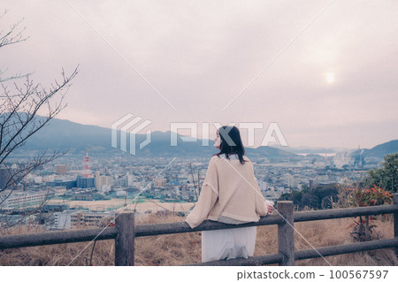 照片中一位女士拿著咖啡杯站在早晨的陽光下，背景是城市 100567597