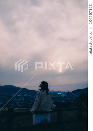 照片中一位女士拿著咖啡杯站在早晨的陽光下，背景是城市 100567598