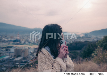 Photo of a woman with a smartphone standing in the morning sun with the city in the background 100567802