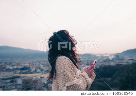 Photo of a woman listening to music with the city in the background in the morning sun Photo of a woman listening to music with the city in the background in the morning sun 100568540