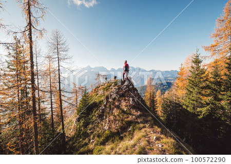 Majestic view of lonley woman hiker standing on top of the hill, autumn forest surrounding her, looking at the view of the mountains in the background 100572290