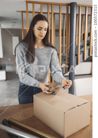 Vertical photo of a woman using tape so seal the cardboard box with Christmas gifts for the family 100572315