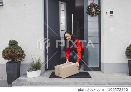 Young woman in red Christmas sweater picking up a cardboard box mailman left out at the front door 100572320