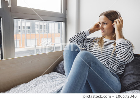 Blonde woman college student relaxing on her bed listening to some music while looking out the window 100572346