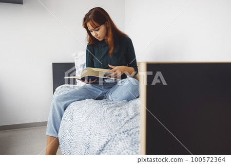 Young woman studying music reading a book on her bed in the dorm room 100572364