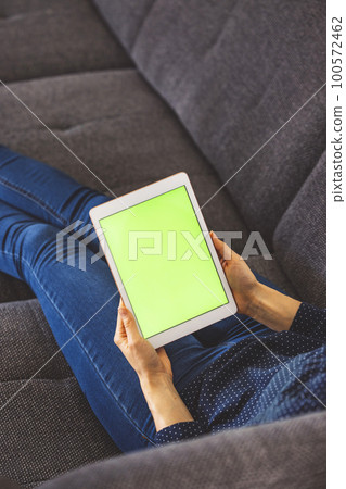 Vertical photo of unrecognizable woman lying on the couch, holding a tablet with green screen in her hands Vertical photo of unrecognizable woman lying on the couch, holding a tablet with green screen in her hands 100572462
