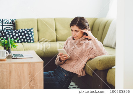 Young woman sitting on the carpet leaning on the couch while looking at her phone 100585896