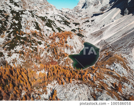 Small emerald green lake in Triglav national park in autumn, occasional trees on the high altitude turning orange in autumn  100586058