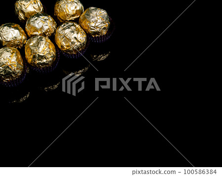 Round chocolate candies in a golden wrapper on a black background. 100586384