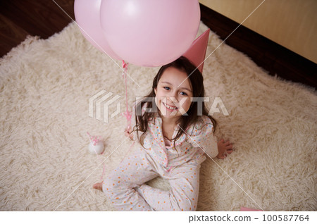 Close-up top view of a lovely 5-6 years old little child, birthday girl in pajamas and party hat, sitting on the carpet near gift box and pink pastel inflatable balloons, smiling looking up at camera 100587764