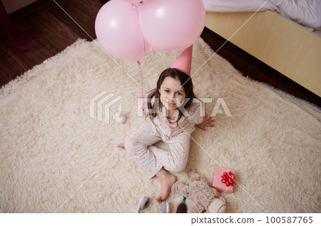 View from above of a charming 5-6 years old little child, birthday girl in stylish pajamas and pink festive hat, sitting on carpet near gift box and helium balloons, smiling looking up at camera View from above of a charming 5-6 years old little child, birthday girl in stylish pajamas and pink festive hat, sitting on carpet near gift box and helium balloons, smiling looking up at camera 100587765