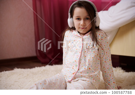 Cute little child girl in stylish comfortable pajamas, enjoying new playlist soundtrack on wireless headphones, looking confidently at camera, sitting on the carpet in her bedroom. Children Lifestyles 100587788