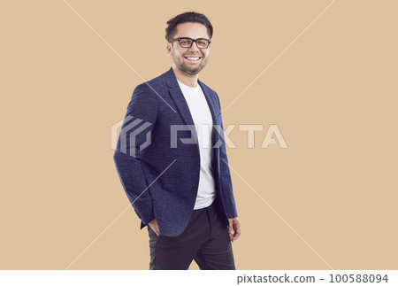 Smiling brunet confident businessman in blue jacket with hand in pocket on beige background. 100588094